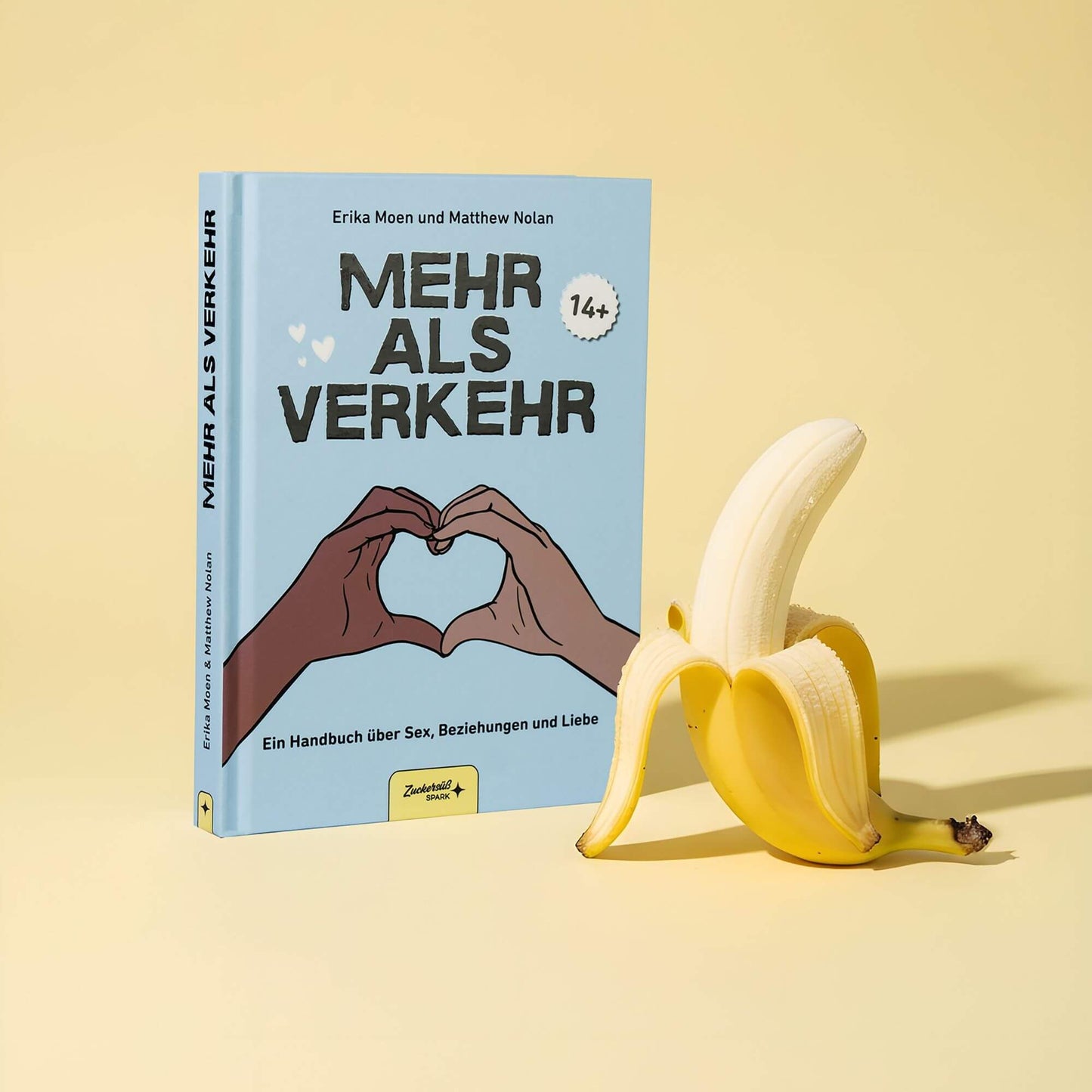 Das Buch Mehr als Verkehr aus dem Zuckersüß Verlag steht aufrecht neben einer geschälten Banane auf gelbem Grund. Das Cover zeigt zwei Hände, die ein Herz bilden, und ist für Leser ab 14 Jahren gedacht.