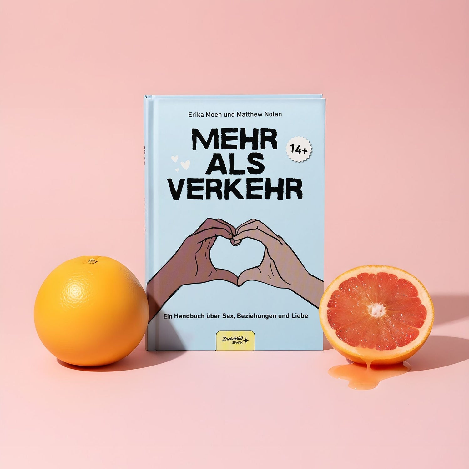 Das Buch "Mehr als Verkehr" vom Zuckersüß Verlag steht aufrecht zwischen einer ganzen und einer halbierten Grapefruit auf rosa Hintergrund. Das Cover zeigt zwei Hände, die ein Herz mit Herzen und deutschem Text bilden.