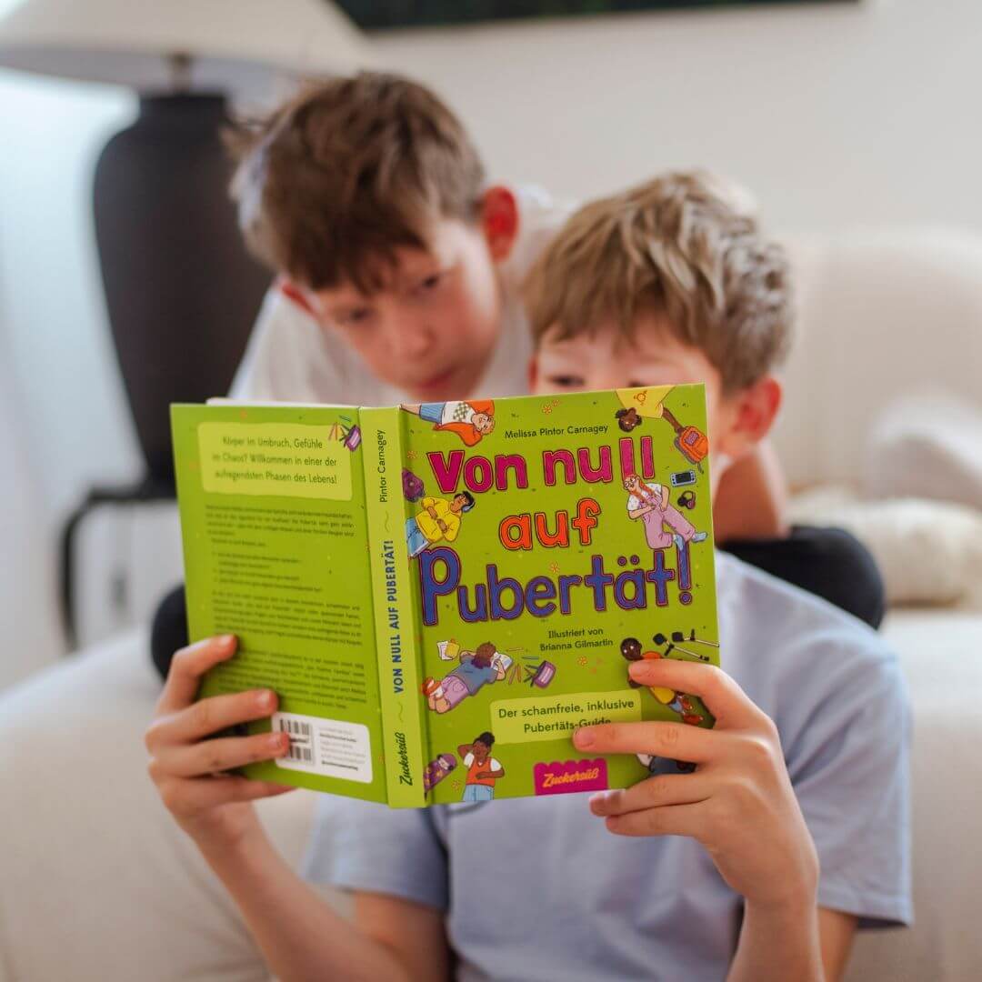 Zwei Jungen sitzen drinnen und beschäftigen sich mit "Von Null auf Pubertät" aus dem Zuckersüß Verlag - ein farbenfrohes deutsches Buch, das sich mit der Pubertät, den körperlichen Veränderungen und der psychischen Gesundheit beschäftigt, wie das verspielte Cover vermuten lässt.