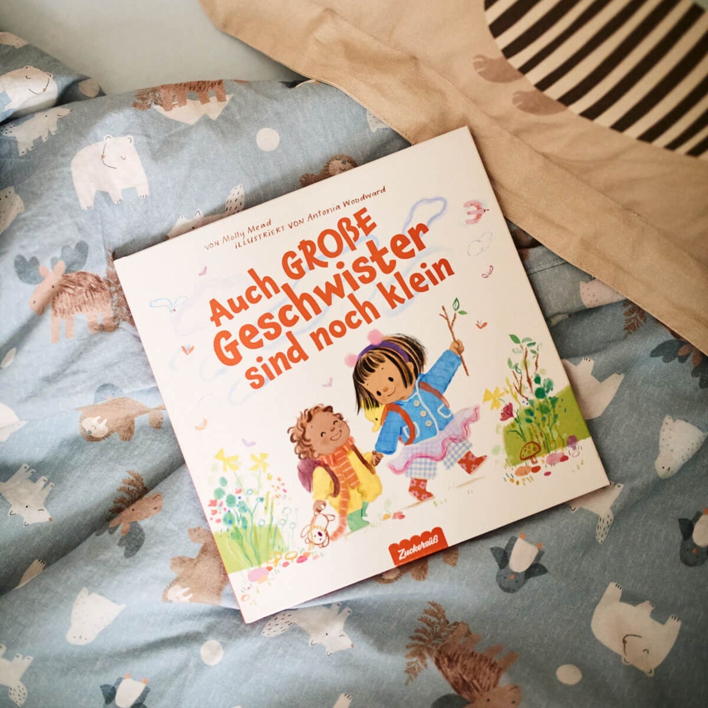 Das deutsche Bilderbuch "Auch große Geschwister sind noch klein" aus dem Zuckersüß Verlag liegt auf einer blauen Tagesdecke mit Tiermotiven; auf dem Cover sind ein Mädchen und ein Junge zu sehen, die draußen spielen und ihre Gefühle erkunden.