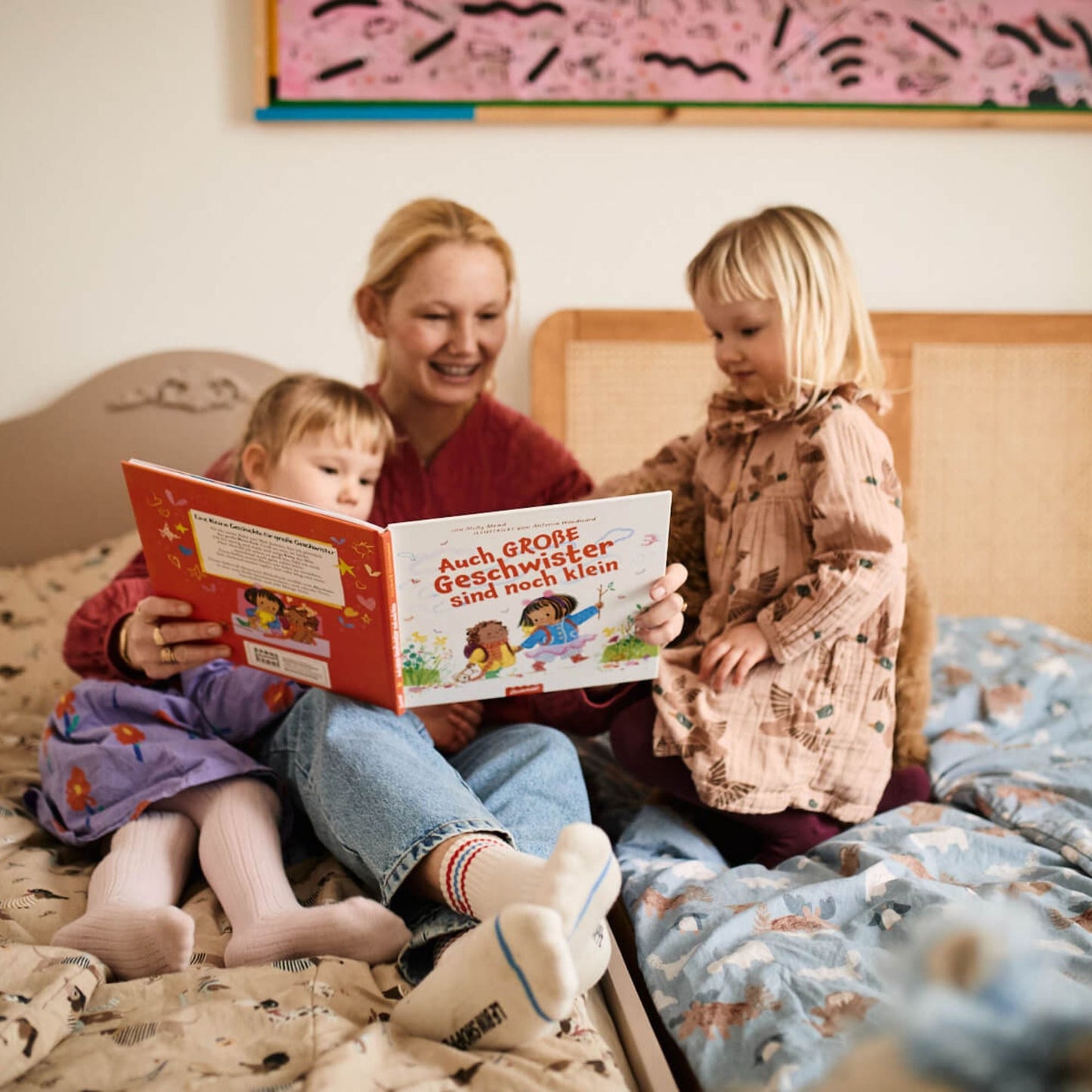 Eine Frau sitzt auf einem Bett und liest zwei jungen Mädchen "Auch große Geschwister sind noch klein" aus dem Zuckersüß Verlag vor. Die Mädchen hören aufmerksam zu - gemeinsam lachen sie und sind neugierig in einer warmen, gemütlichen Schlafzimmeratmosphäre.